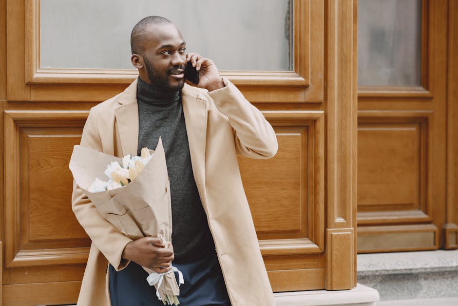 A man with dark skin and a neatly groomed beard is holding a bouquet of fresh flowers wrapped in brown paper while standing outside a building with wooden paneling and large glass windows. He is weari