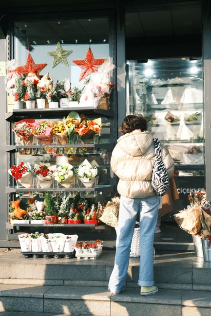 A woman with curly hair, wearing a beige puffer jacket and light blue jeans, stands outside a flower shop with large glass windows. She is holding a striped handbag and several paper shopping bags. Th