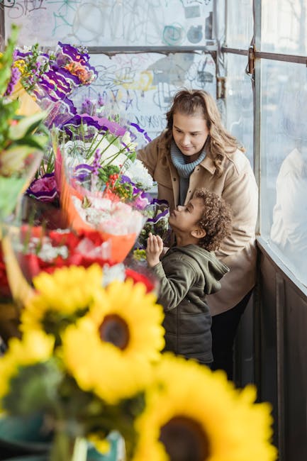 A woman and a young child are inside a greenhouse or flower shop, surrounded by a vibrant display of fresh floral arrangements. The woman has long, wavy brown hair and is wearing a beige coat and a li