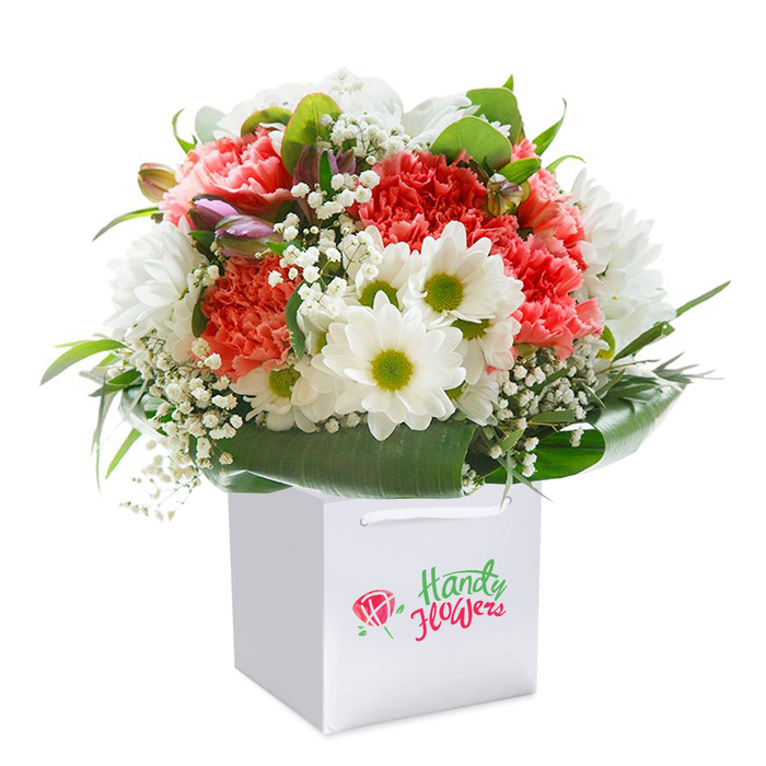 Vivid coral and red carnations burst with energy in this carefully crafted bouquet, rising from a crisp square gift bag that hints of celebration and care. The carnations' plush, multi-layered petals bring depth and texture, while bright white daisies with sunlit yellow-green centers add a joyful, welcoming air. The entire composition rests atop a luxuriant layer of deep green foliage, whose glossy surfaces cradle the central blooms much like the leafy shelters spotted throughout Norwood Junction's quieter corners. Misty, cloud-like baby's breath offers a delicate lacework between flowers, and subtle purple-tinged buds provide gentle contrast, elevating the overall design with a sense of curiosity and movement. The white gift bag itself, practical and elegant with its secure ribbon handles, is arranged to suggest both ease of transport and the excitement of presenting a meaningful gift-perfect for birthdays, anniversaries, or simply sending kind wishes across South Norwood. Shot against a flawless white background, the bouquet stands out for its contemporary feel, balancing abundant colour with a measured, florist's touch. The arrangement's fresh, lively presence evokes the unique vibrancy found around South Norwood's lively markets, and the bouquet's ready-to-gift aesthetic captures the essence of sincere, thoughtful flower delivery within the local community.