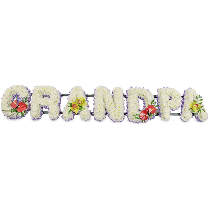 This image shows an elegant floral tribute spelling out "GRANDPA" in bold, upright capital letters, displayed in a single line against a plain white background so every bloom is clearly visible. Each letter is built from compact clusters of small white chrysanthemums, placed so closely together that they form a velvety blanket of petals, creating a soft, cushion-like surface. The pure white is gently framed by a neat edging of pale lavender ribbon running along the base of each letter, introducing a cool, pastel tone that adds contrast and a sense of refined detail without overwhelming the design. Discreet dark green rails link the letters, giving the tribute stability while remaining visually unobtrusive. Splashes of richer colour appear in carefully positioned sprays: the "G" on the left and the "D" near the centre carry bold groups of deep crimson-red anthuriums, their distinctive heart-shaped, glossy spathes and pale yellow spadices nestled into dark green foliage and tiny hints of purple filler. The first "A" and the final "A" on the right are graced with clusters of yellow-green orchids, their slim petals arching delicately among fresh leaves, offering a touch of gentle brightness and sophistication. The overall look is dignified and deeply personal, the kind of bespoke "Grandpa" tribute a South Norwood family might arrange for a funeral service near Woodside Green or a burial in a nearby cemetery. It conveys love, gratitude, and remembrance through thoughtful craftsmanship and a tranquil blend of white, red, and soft green tones.